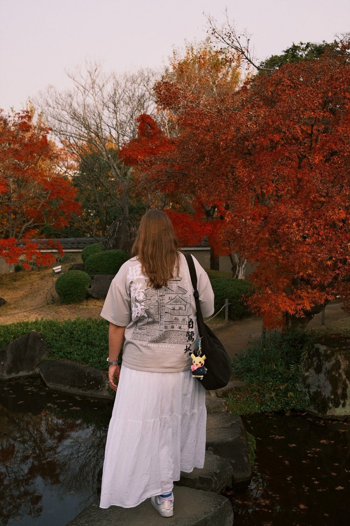 A woman in an INCIDENT T-shirt and a long white skirt stands under red maples in a Japanese garden.