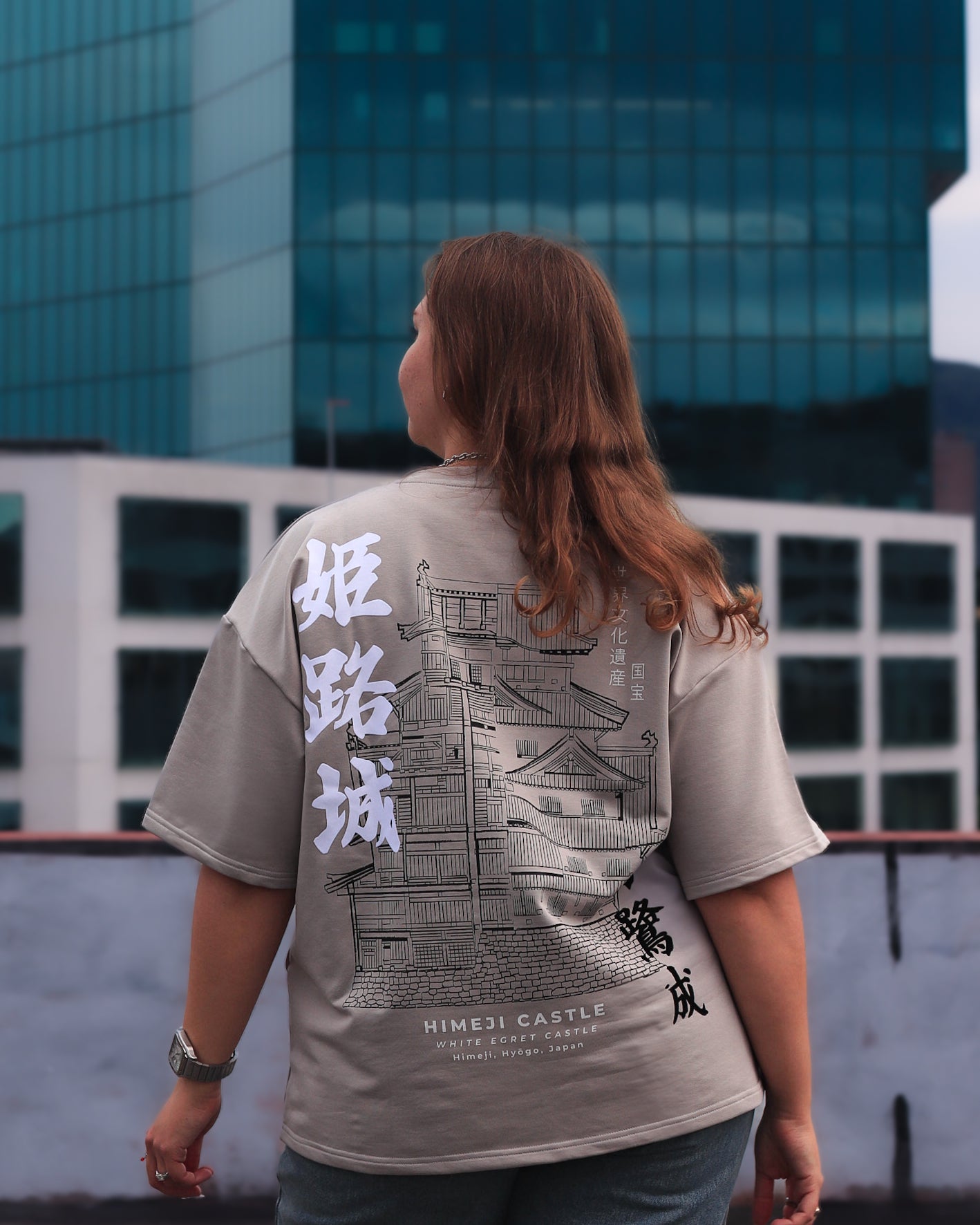 Person wearing a gray t-shirt with architectural design and text, standing in front of a modern building.