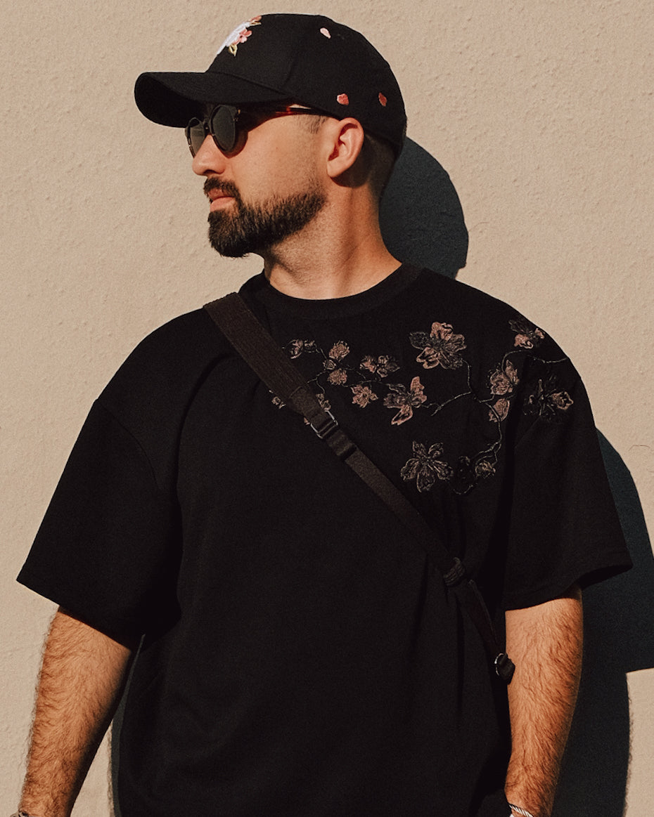 Man wearing a black cap and black t-shirt with floral embroidery against a beige wall.