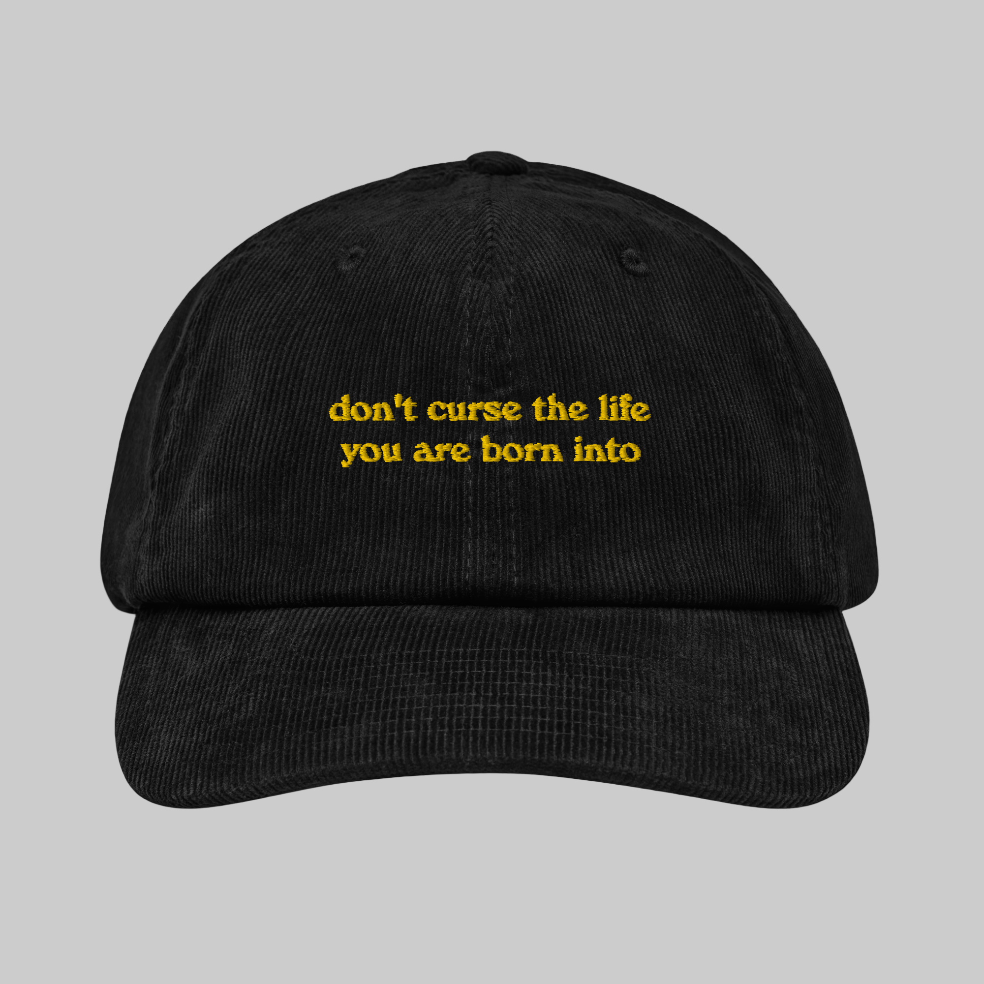 A black corduroy 6-panel cap with an adjustable strap, featuring yellow text on the front that reads 'don't curse the life you are born into'.