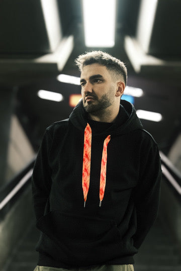 A person wearing a black hoodie with a distinctive stripe pattern and a dragon design on the back standing in a dimly lit space.