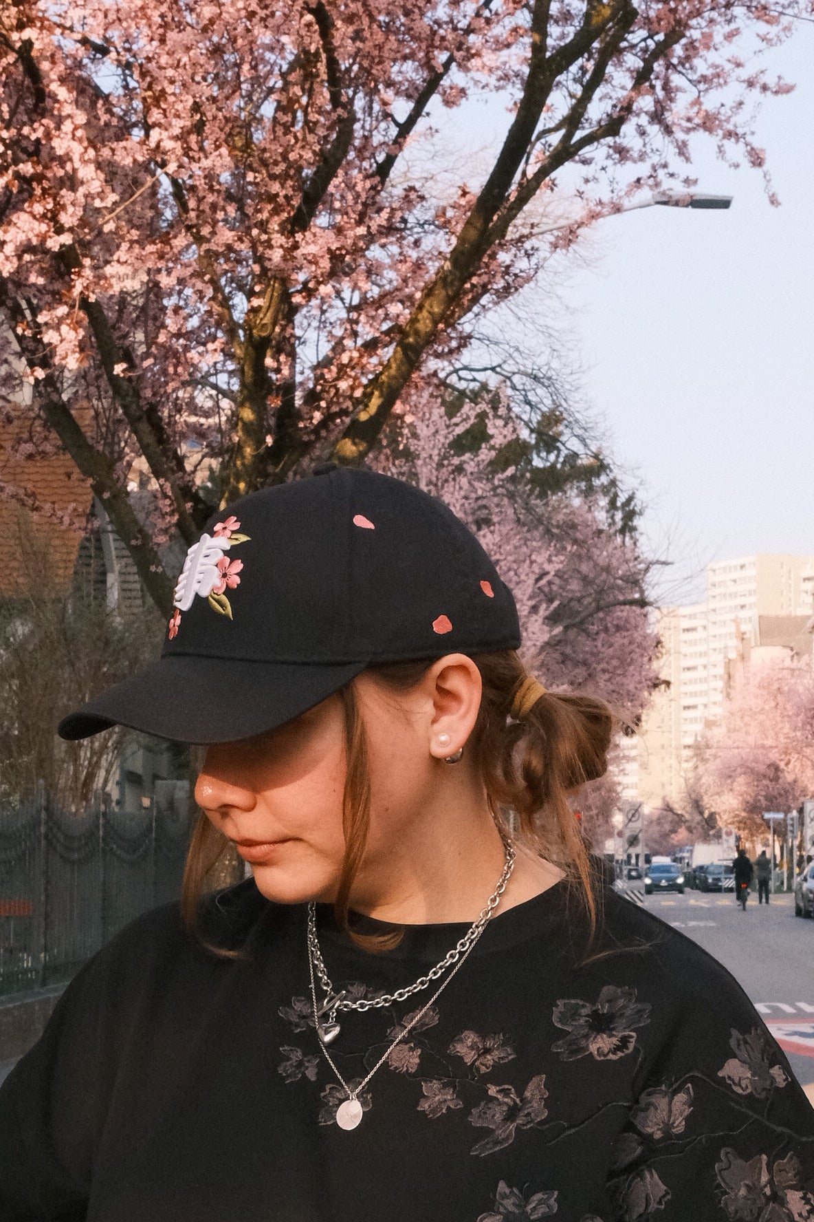 Woman wearing a black INCIDENT cherry blossom cap and floral top, with cherry blossom trees in the background