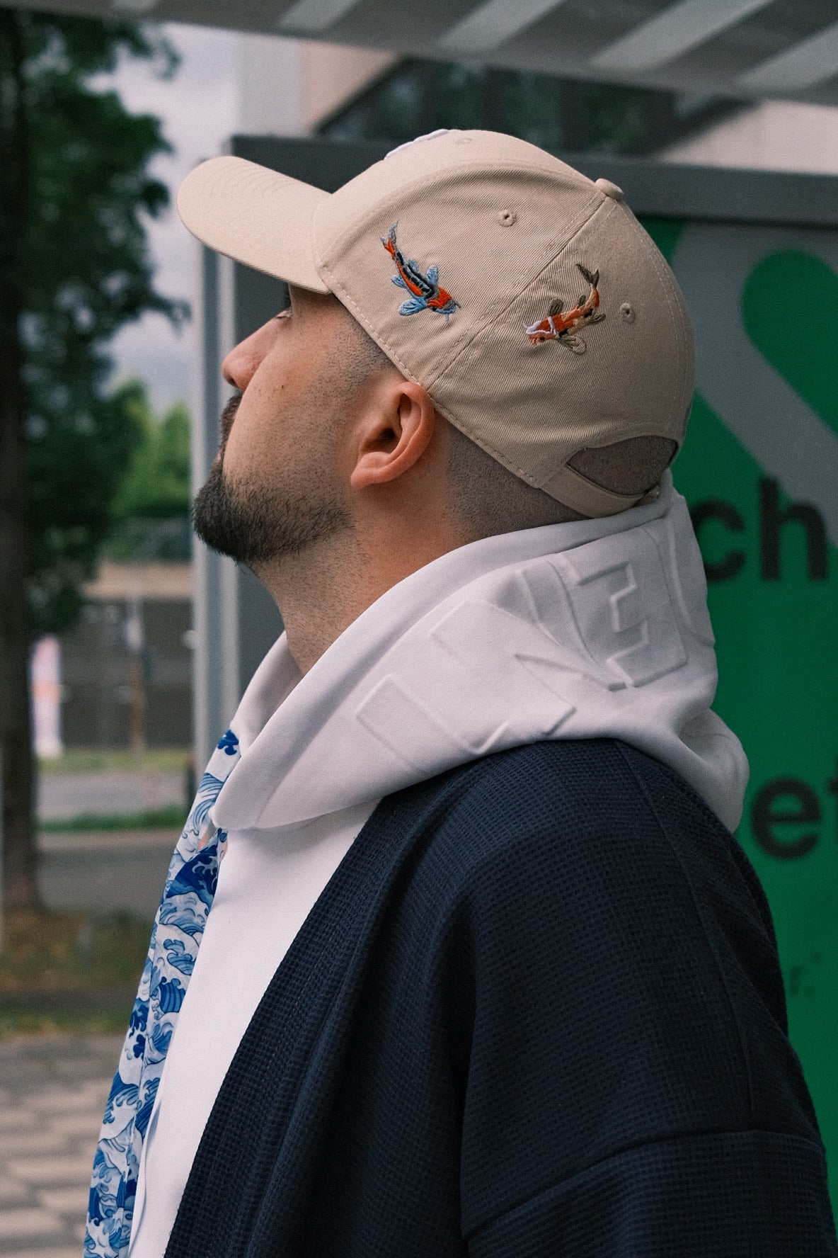 Man wearing a beige INCIDENT cap with embroidered koi, looking away from the camera.