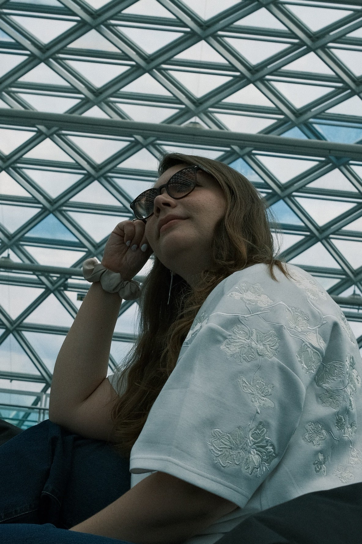 Woman wearing sunglasses and a white embroidered INCIDENT T-Shirt, sitting inside a geometric glass structure.
