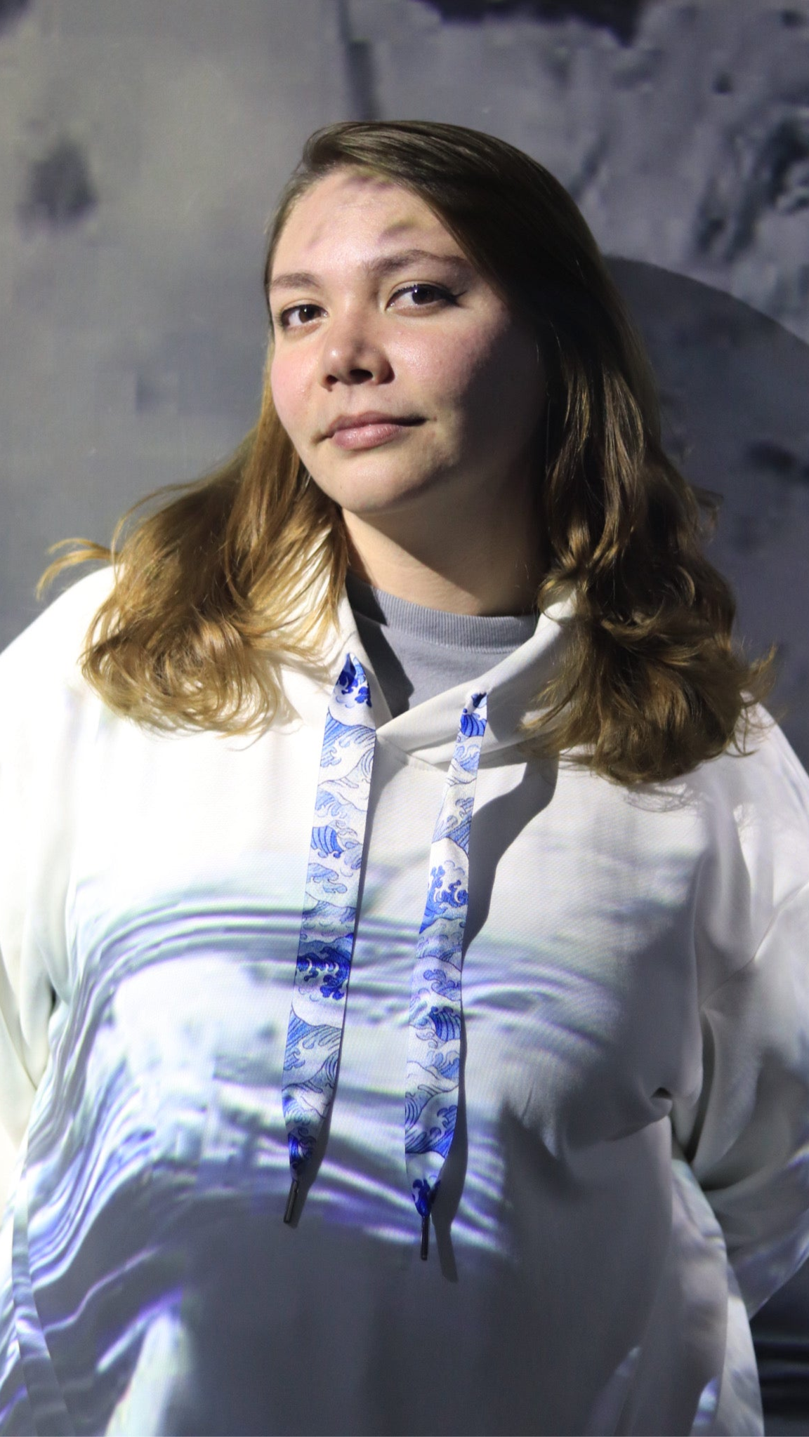 Person wearing a white hoodie with a blue and purple design, standing against a textured gray background.